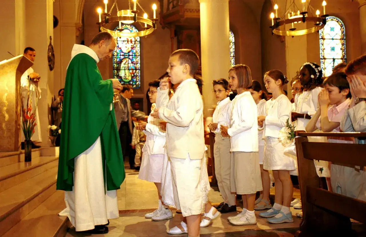 Cérémonie religieuse : un prêtre distribue la communion à des enfants. Peut-on être marraine à 11 ans ? La question se pose devant ces jeunes visages.