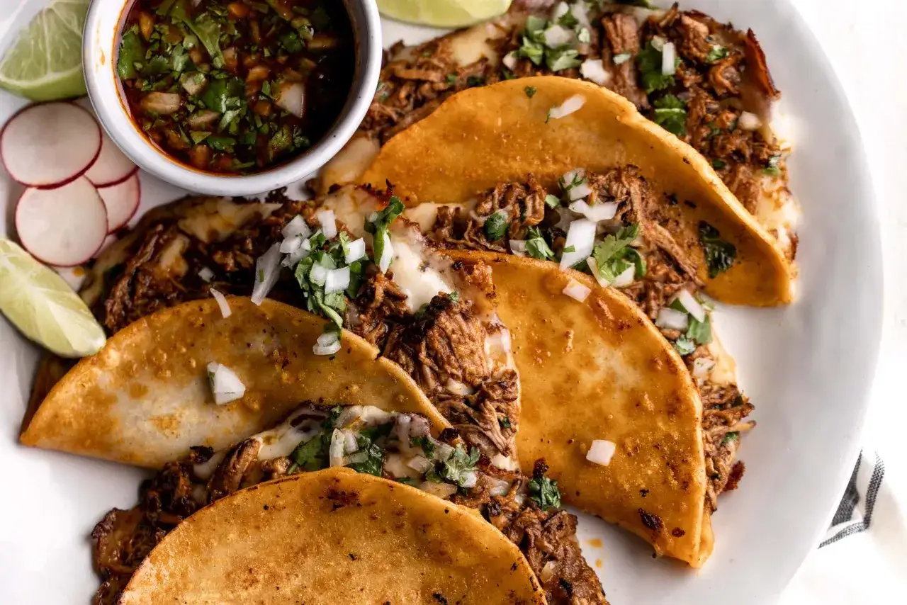 En tallrik med gyllene birria tacos, fyllda med mört kött och ost, toppade med lök och koriander. Serveras med en skål consommé, limeklyftor och skivad rättika.