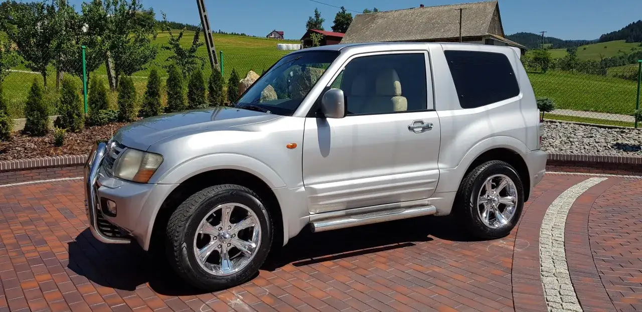 Srebrne Mitsubishi Pajero 3 na podjeździe z kostki brukowej, w tle zielone wzgórza i domy.