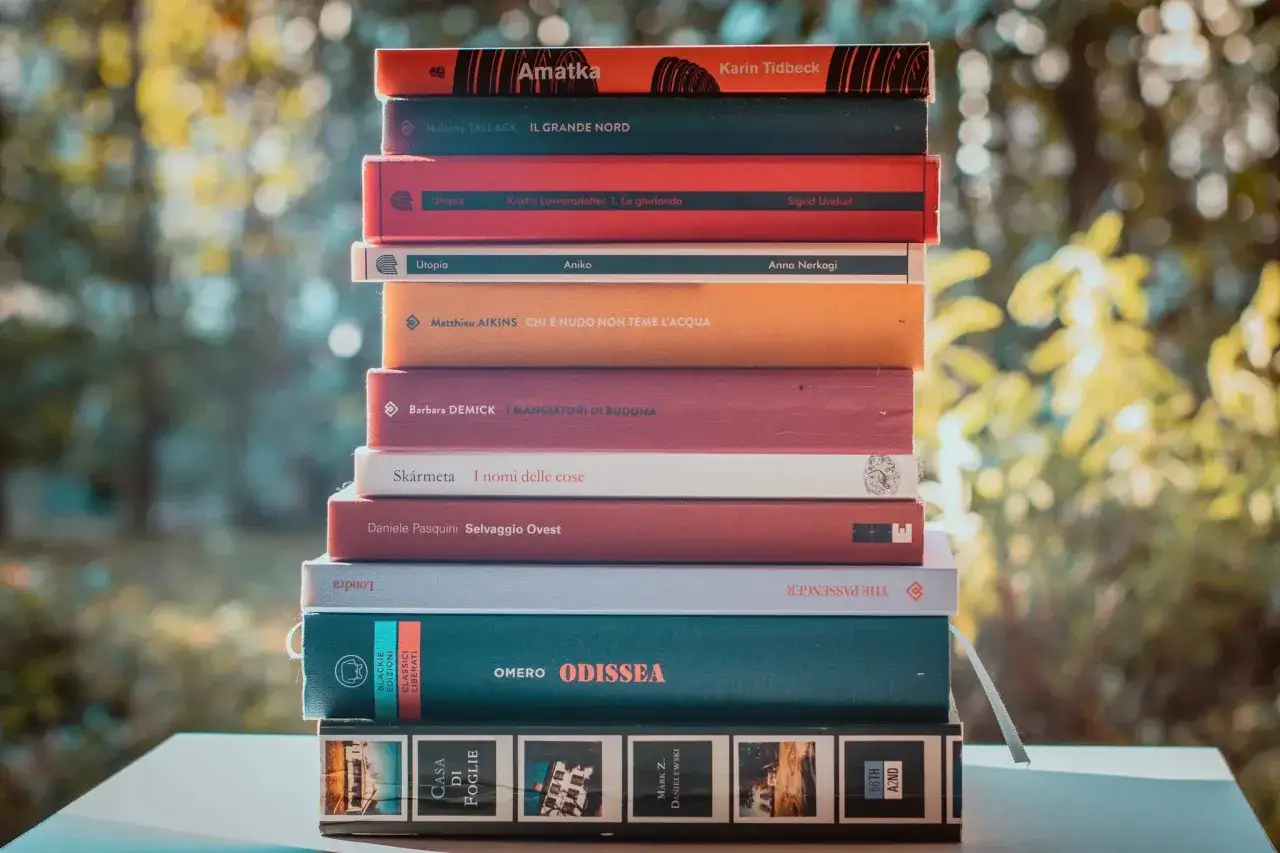 Una pila di bei libri da leggere, con titoli in italiano e inglese, su uno sfondo sfocato di alberi verdi.