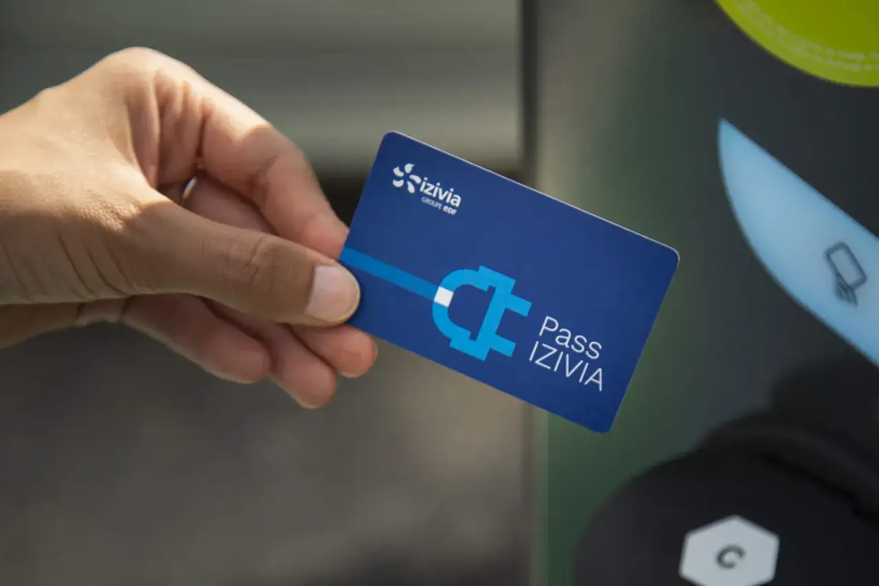 Une main tient une carte pour recharger voiture &eacute;lectrique, le Pass IZIIVIA, devant une borne de recharge.