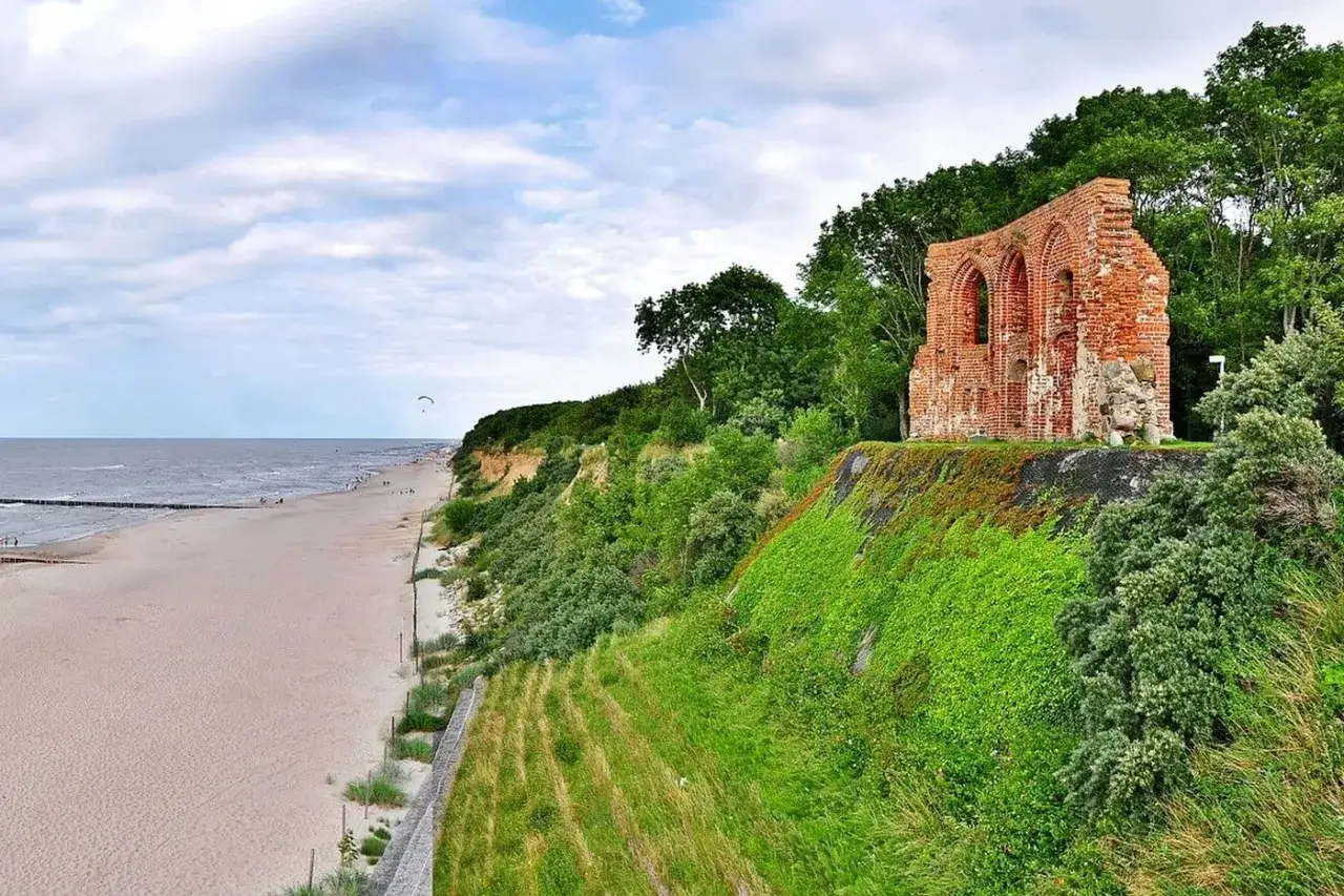 Ruiny ceglanej budowli na zielonym wzgórzu nad piaszczystą plażą i morzem. Klif nad morzem zachwyca widokiem.