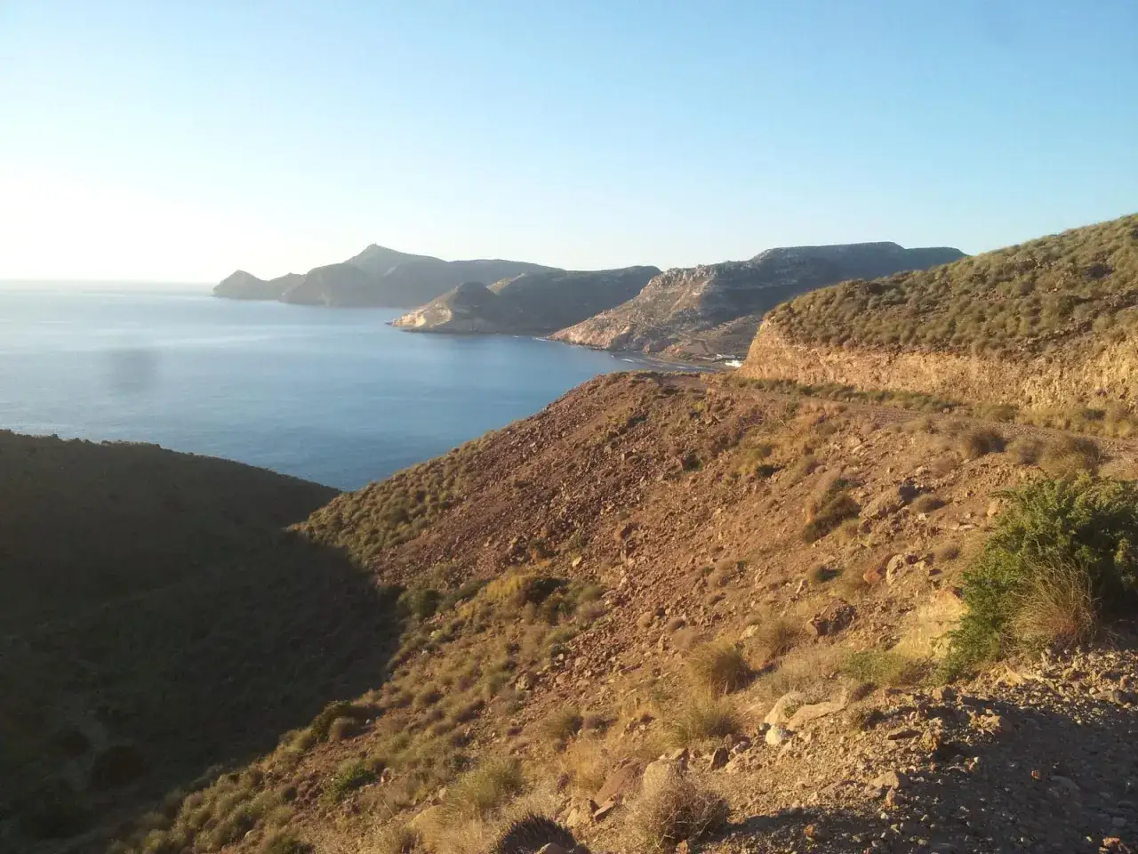 Paisaje rocoso y árido con vegetación escasa que desciende hacia el mar azul. Se vislumbran montañas y la costa de cala San Pedro al fondo.