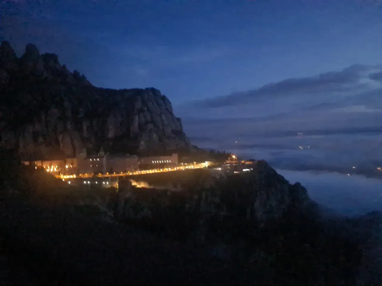 Creu de Sant Miquel il&middot;luminada al capvespre, amb el monestir aferrat a la muntanya i boira al fons.