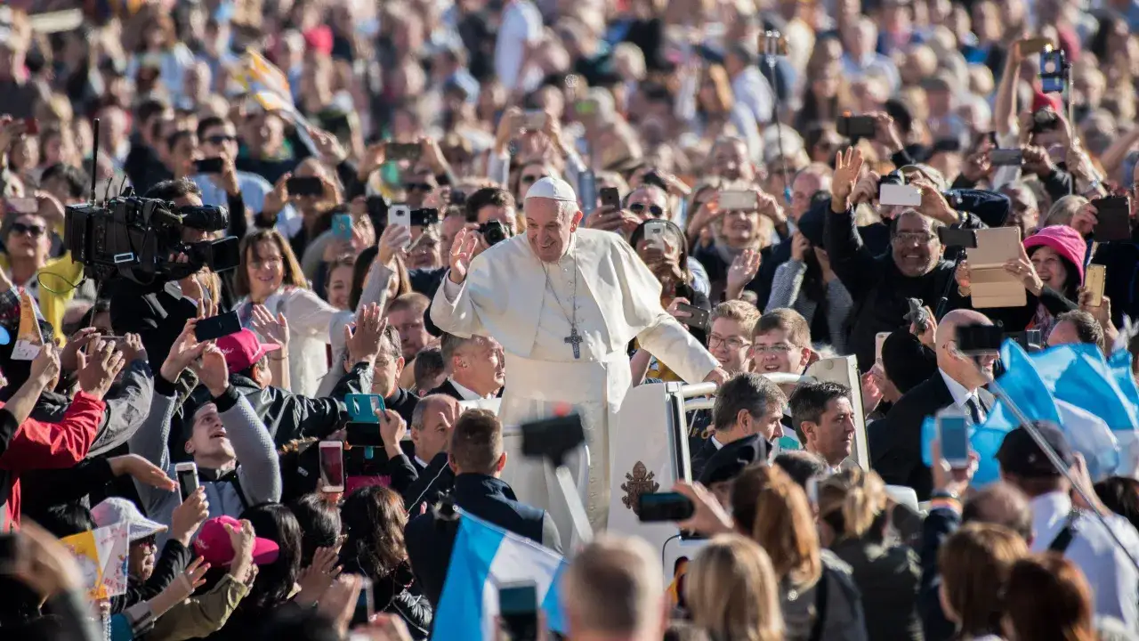 Pope Francis waves to a cheering crowd in Rome, with many people holding up phones to capture the moment.