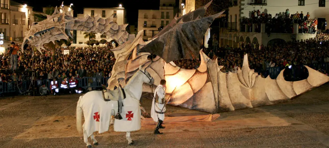 Caballero blanco en caballo, frente a un dragón gigante, en una celebración del **San Jorge día**. Multitud observa el espectáculo nocturno.