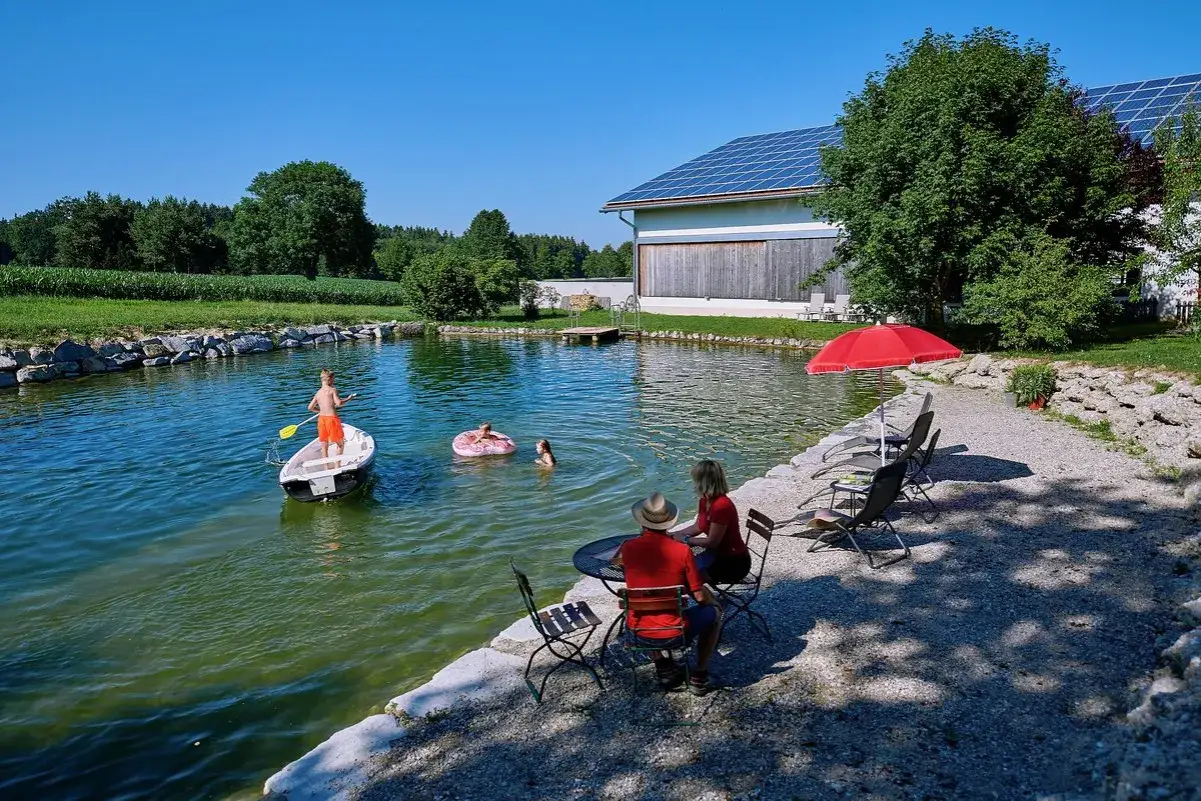 Familie genießt Ferien auf dem Bauernhof Bayern: Kinder planschen im Teich, während Eltern am Ufer entspannen.