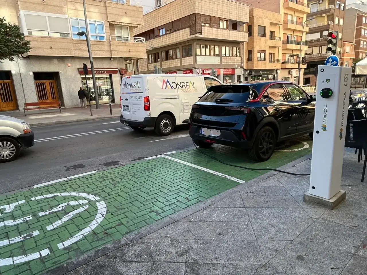 Coche el&eacute;ctrico negro cargando en la calle, en una zona verde con se&ntilde;al de carga. As&iacute; se ve como cargar coche electrico en la calle.