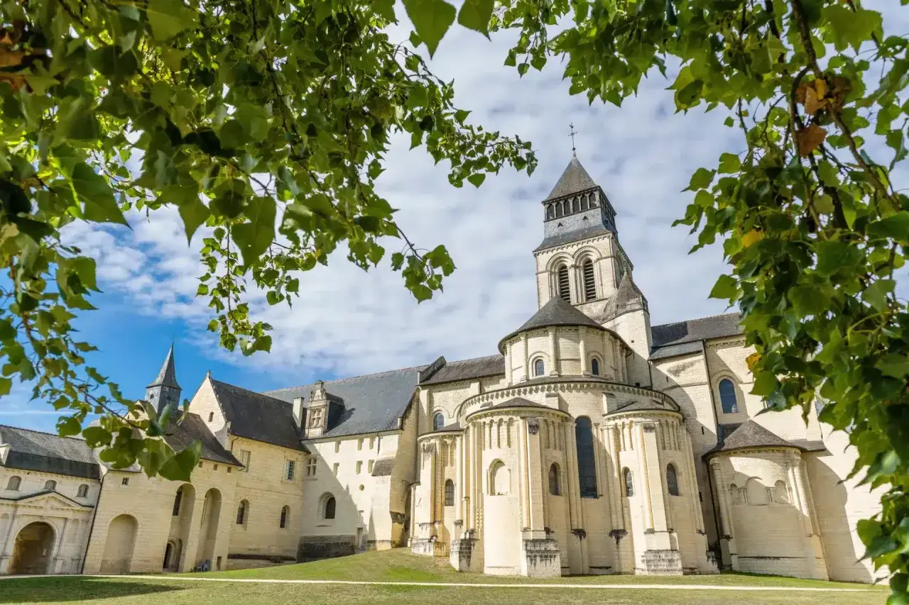 Le majestueux **château de Fontevraud**, avec sa tour imposante et ses absides romanes, se dresse sous un ciel bleu parsemé de nuages, encadré par le feuillage verdoyant des arbres.