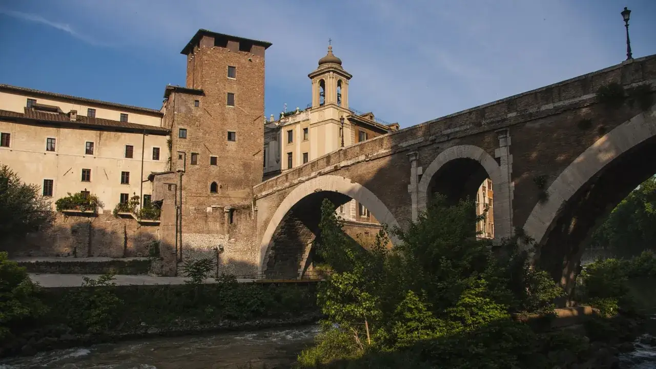 El hist&oacute;rico puente Fabricio cruza el r&iacute;o T&iacute;ber, conectando la isla Tiberina con la orilla. Edificios antiguos y vegetaci&oacute;n exuberante lo rodean.