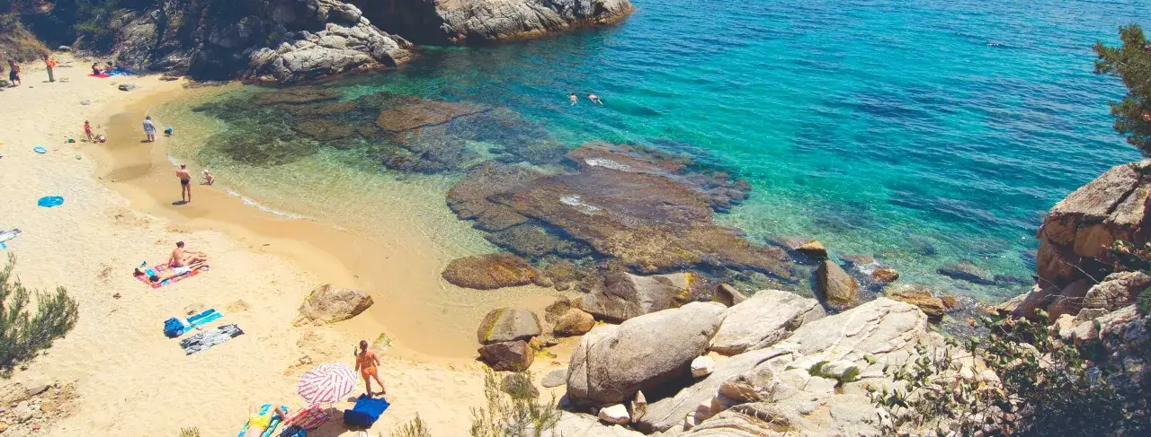 Una cala en la Costa Brava con arena dorada, aguas turquesas y rocas. Gente disfrutando del sol y el mar.