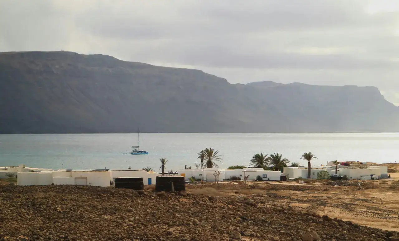 Paisaje de los Hervideros Lanzarote con casas blancas, palmeras y un catamarán navegando en el mar azul bajo una montaña imponente.