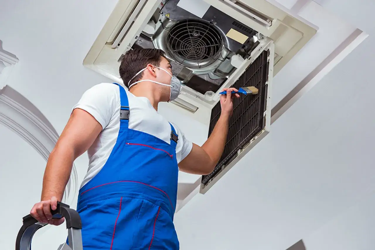 T&eacute;cnico con mascarilla realiza mantenimiento de aire acondicionado, limpiando el filtro con un cepillo.