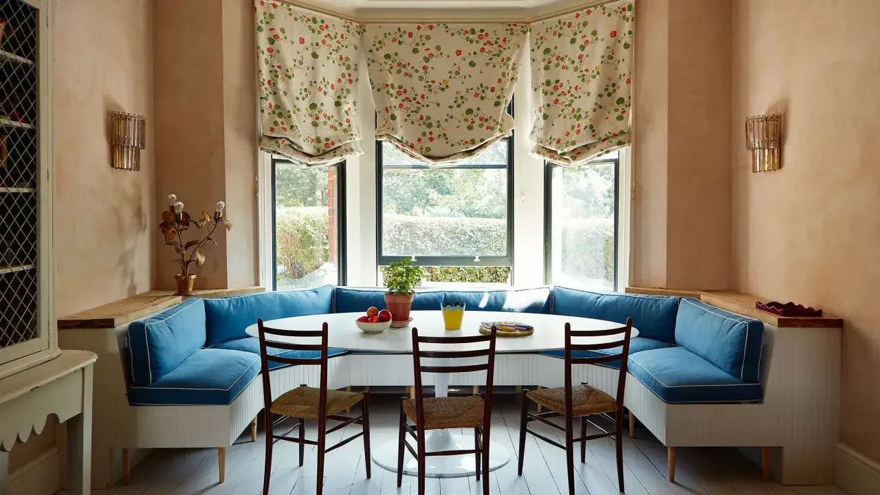A cozy breakfast nook with blue banquette seating, a white oval table set with fruit and a yellow vase, and floral Roman shades. Simple pleasures abound in this inviting space.