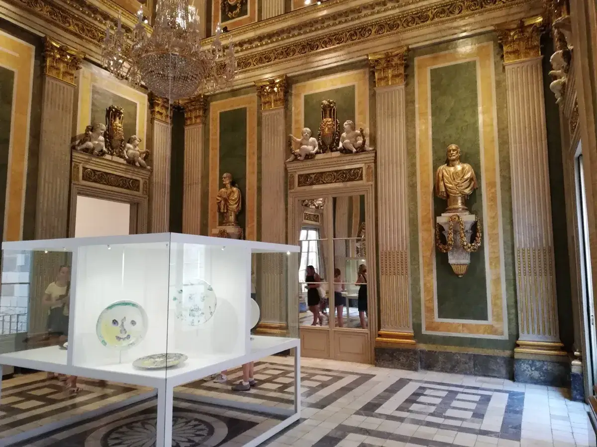 Wnętrze muzeum Picassa w Paryżu. W szklanej gablocie wystawiono kolorowe talerze. Złote zdobienia i kryształowy żyrandol dodają blasku.