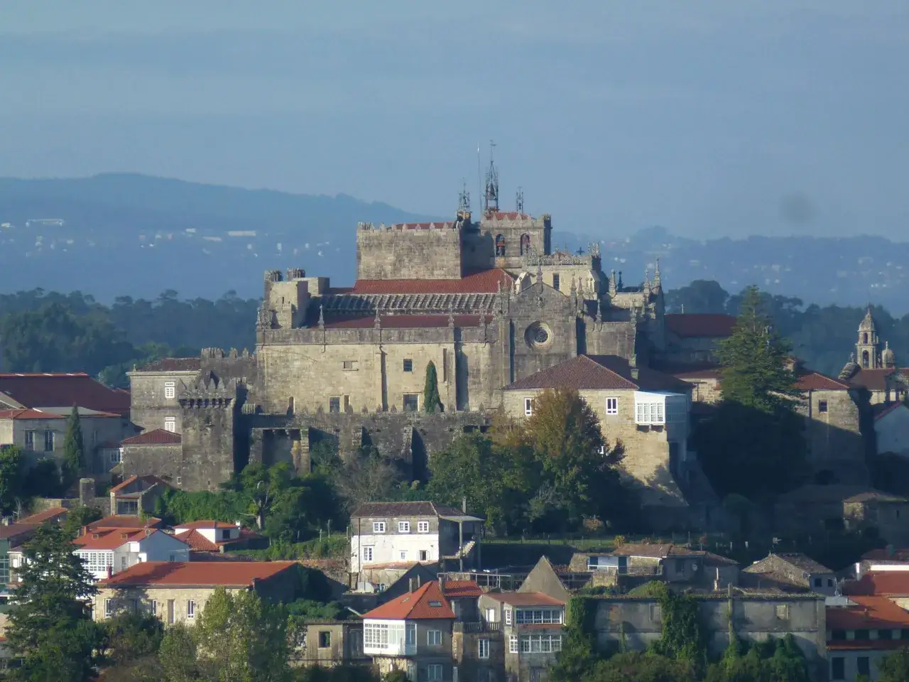 La imponente catedral de Tui se alza sobre un paisaje de tejados rojos y vegetación, con colinas neblinosas al fondo.
