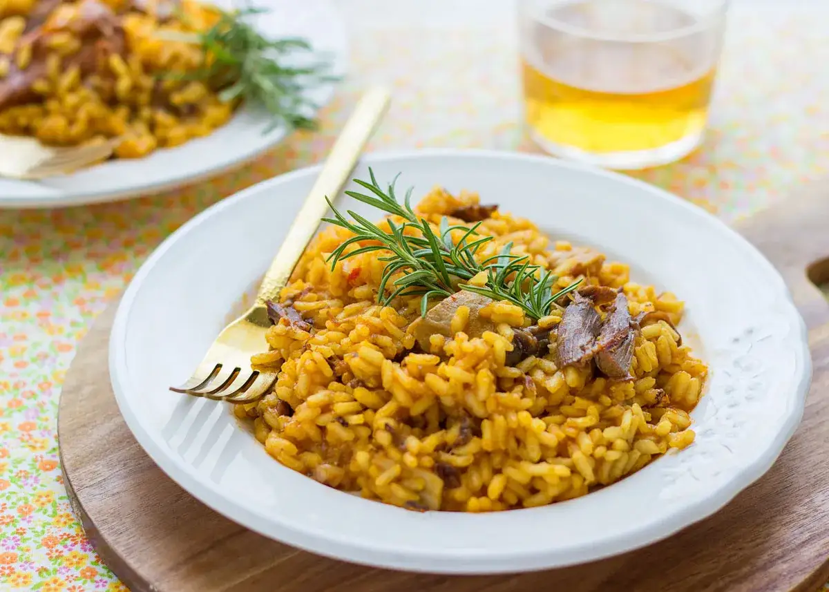 Un plato de arroz con secreto, adornado con romero fresco. El arroz tiene un color dorado intenso y se ve jugoso, con trozos de carne tierna.
