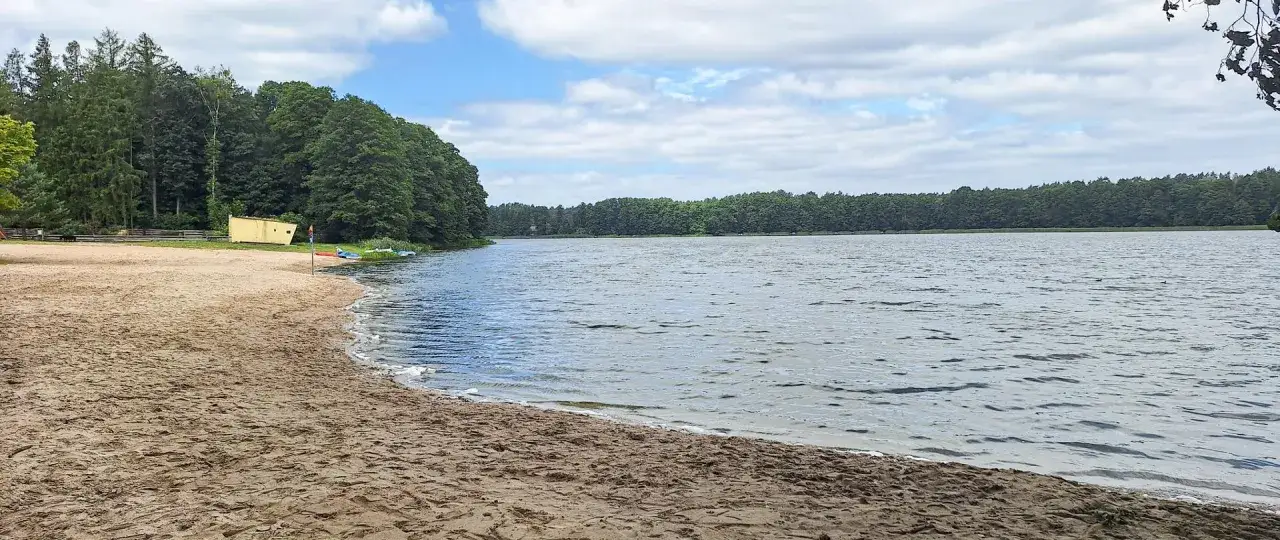 Plaża nad jeziorem, las w tle. W oddali widać żółty budynek, a na brzegu kajaki. Idealne miejsce na wypoczynek w stylu krzyż wielkopolski.