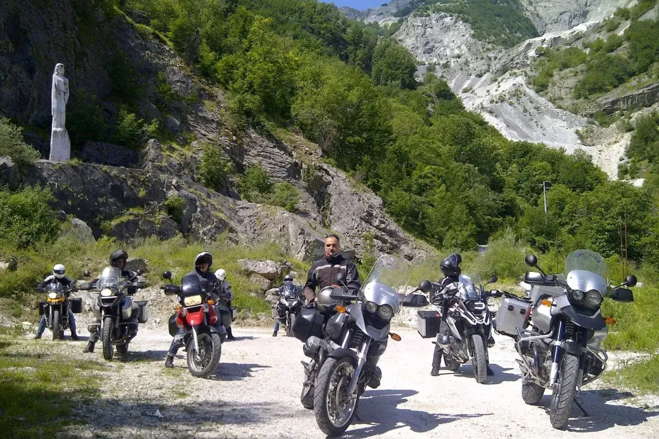 Gruppo di motociclisti in sosta su una strada sterrata, con una statua bianca su un fianco della montagna. Il passo del vestito è ben visibile sui motociclisti.