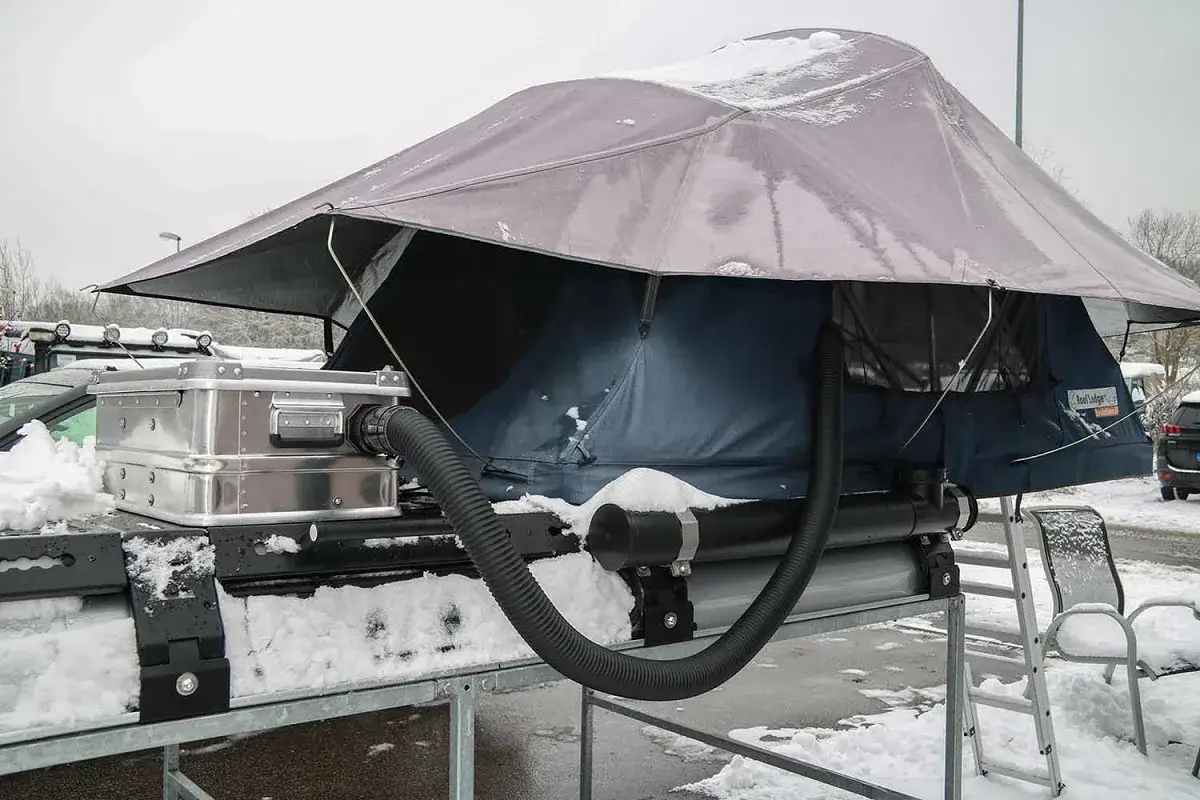 Dachzelt mit Heizung und Schlauchanschluss, bereit für winter camping. Schnee bedeckt die Ausrüstung.