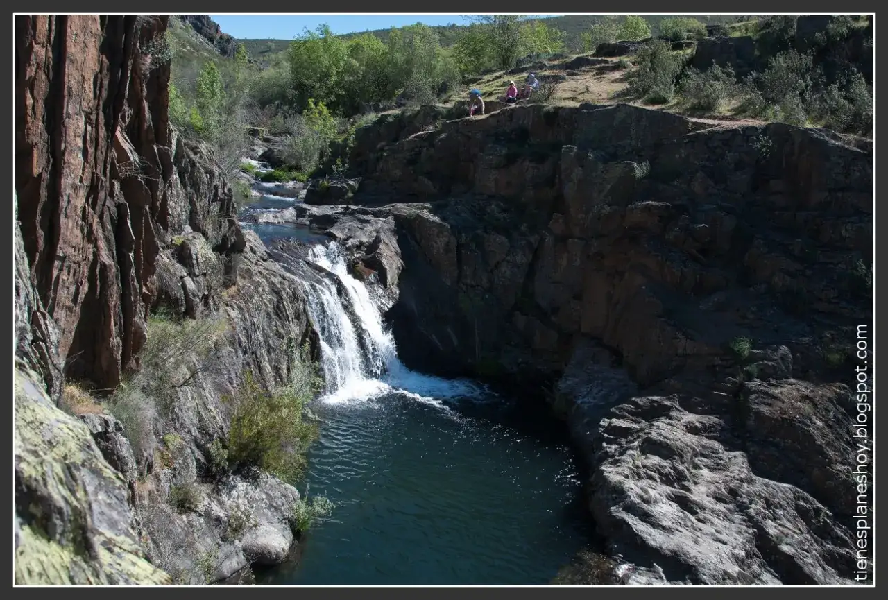 La cascada del aljibe desciende entre rocas y vegetaci&oacute;n hacia una poza de aguas oscuras. Personas observan desde la cima.