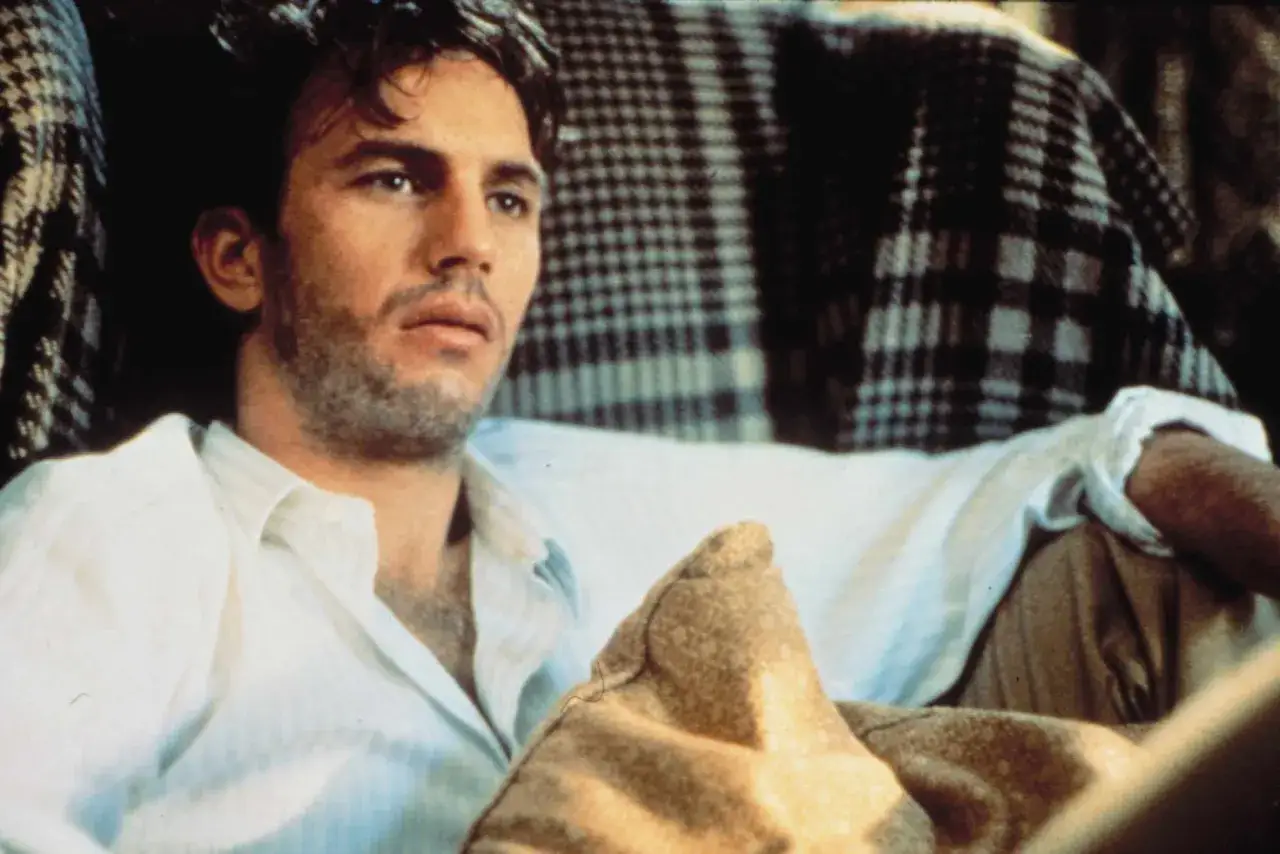 Kevin Costner jung, nachdenklich in einem wei&szlig;en Hemd, umgeben von karierten Decken.