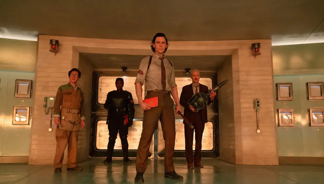 Die Besetzung von Loki steht im Raum. Loki hält ein Buch, ein Mann im schwarzen Anzug eine Waffe.