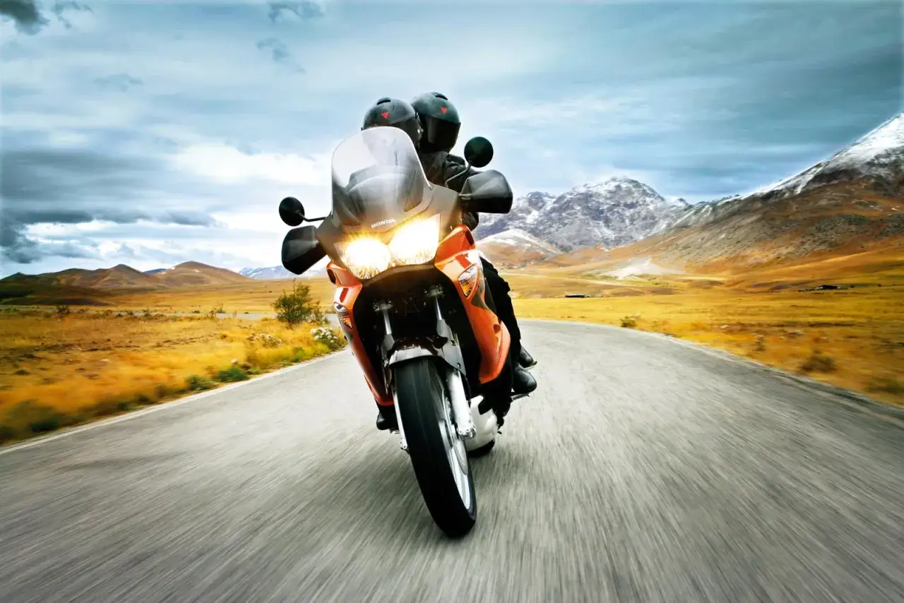 Dwaj motocykliści na pomarańczowym Honda Varadero pędzą krętą drogą przez pustynny krajobraz z ośnieżonymi górami w tle.