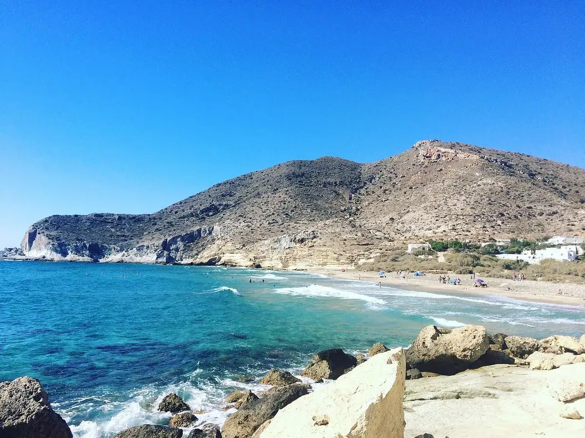 Aguas turquesas bañan la **cala del Plomo**, con gente disfrutando en la playa y montañas áridas de fondo bajo un cielo azul.