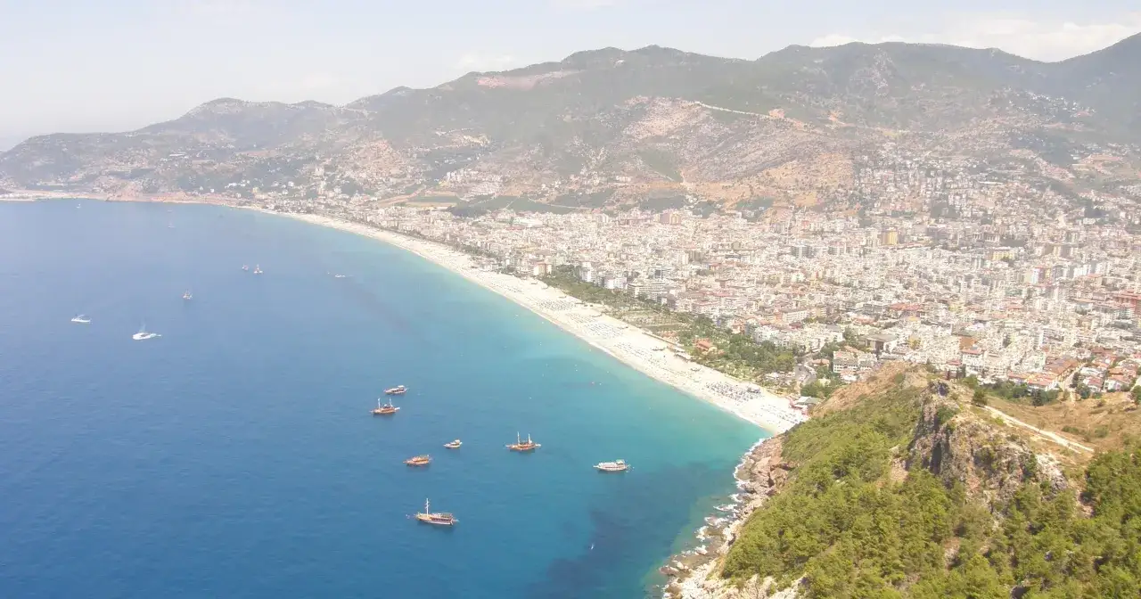Widok na wybrzeże Alanyi z górami w tle. Widać plażę, morze z łodziami i miasto. To wspaniałe alanya atrakcje.