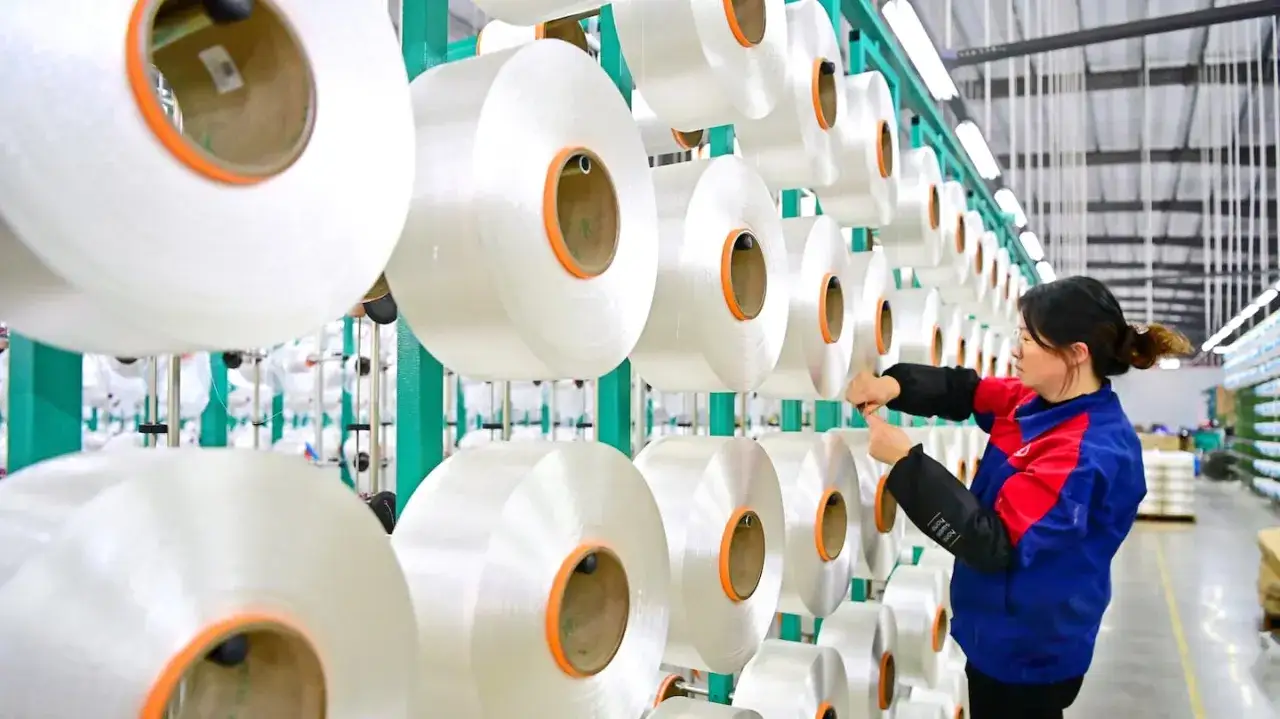 En kvinna arbetar i en fabrik d&auml;r stora rullar med garn h&auml;nger p&aring; st&auml;llningar. Detta &auml;r en del av hur tillverkas polyester, en process som ger oss m&aring;nga av v&aring;ra textilier.