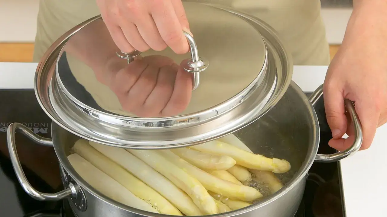 H&auml;nde heben den Deckel eines Topfes, in dem wei&szlig;er Spargel kocht.