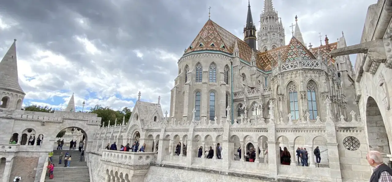 Budynek z kolorowym dachem w Budapeszcie, przypominający zamek. Turyści zwiedzają, myśląc o tym, ile taka waluta w Budapeszcie kosztuje.