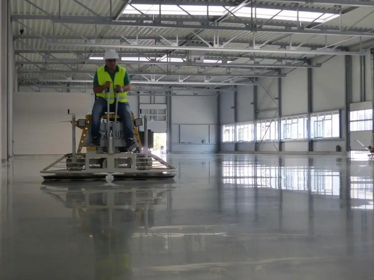 Trabajador con chaleco reflectante operando una m&aacute;quina para alisar un brillante pavimento industrial.