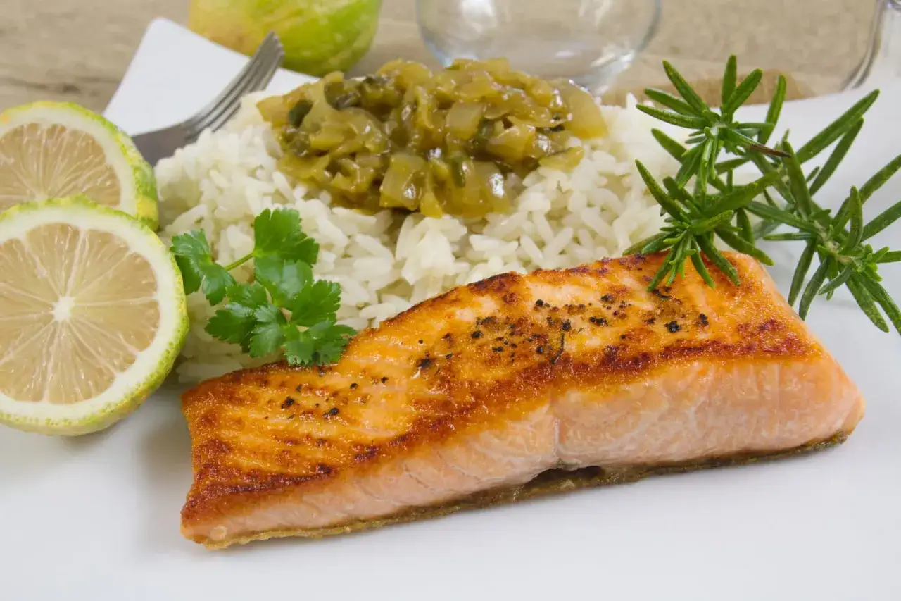 Un pavé de saumon grillé, servi avec du riz blanc, une garniture verte et des quartiers de citron.