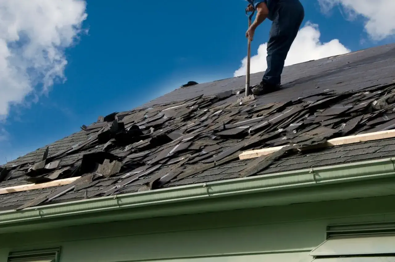Worker on a damaged roof, using a shovel to remove shingles. This shows a step in how to dispose of shingles after a storm.