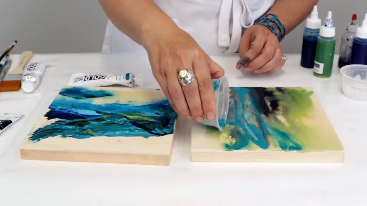 Une main verse un liquide bleu-vert sur une toile, créant des effets abstraits. L'artiste semble mélanger peinture acrylique et résine pour un rendu unique.