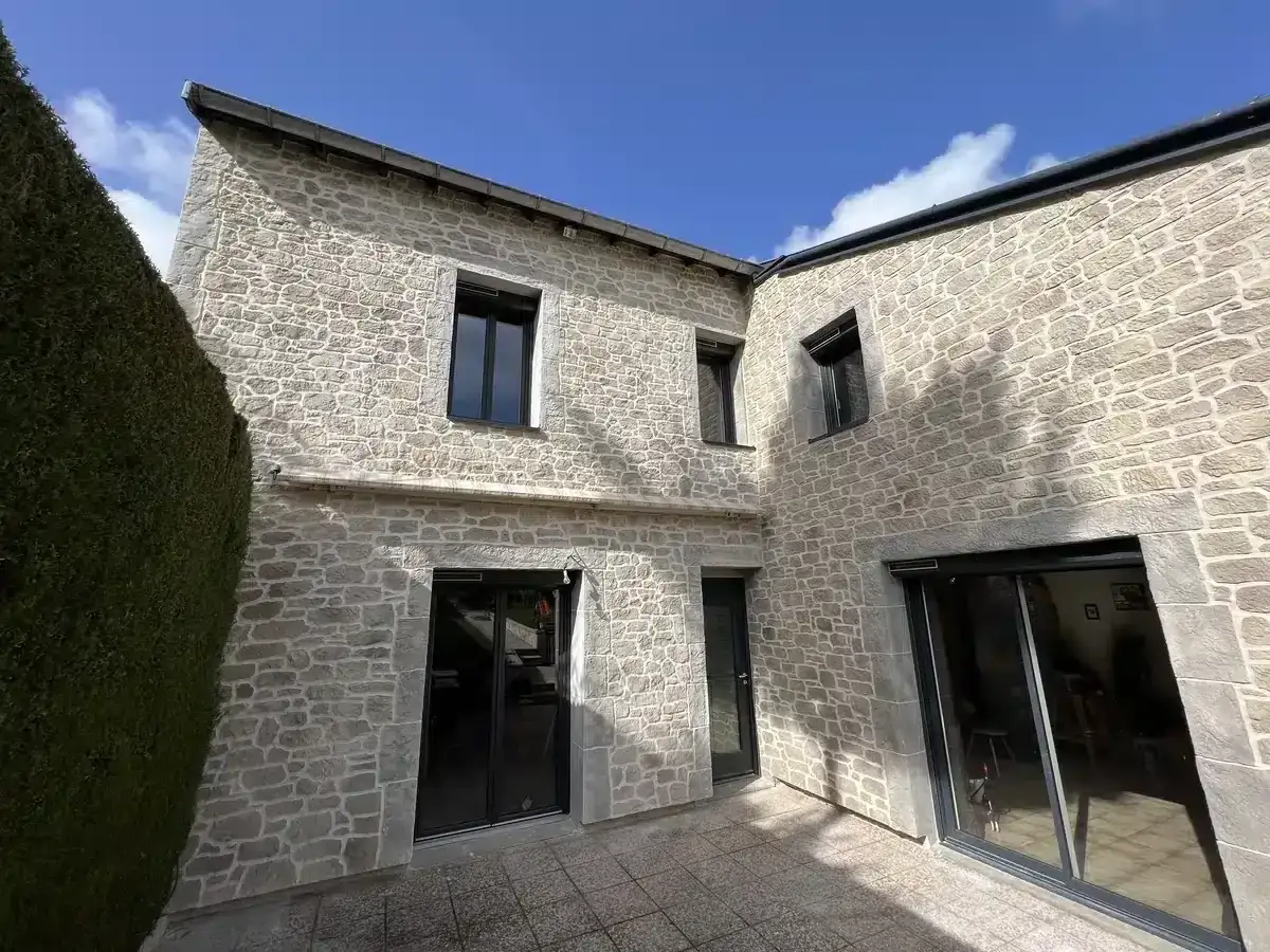 Maison en pierre avec un mur ext&eacute;rieur irr&eacute;gulier, une haie verte luxuriante et un ciel bleu clair.