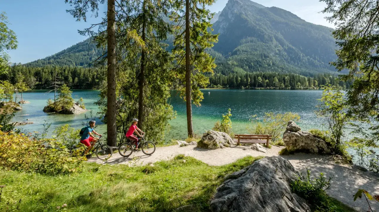 Two cyclists enjoy scenic bike tours in Germany along a crystal-clear lake, with a majestic mountain backdrop.