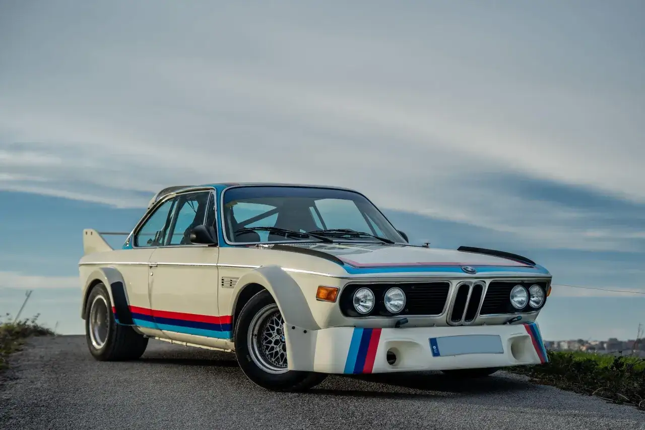 Klasyczne BMW E9 w wyścigowym malowaniu z pasami M. Biały lakier, szerokie nadkola i spojlery podkreślają sportowy charakter.