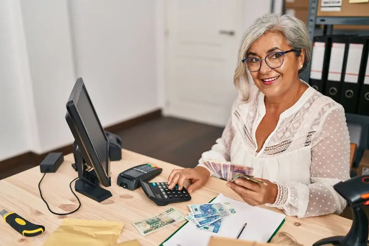 Uśmiechnięta kobieta liczy pieniądze, przykład uzasadnienia nagrody dla pracownika. Na biurku komputer, terminal płatniczy, kalkulator i polskie banknoty.