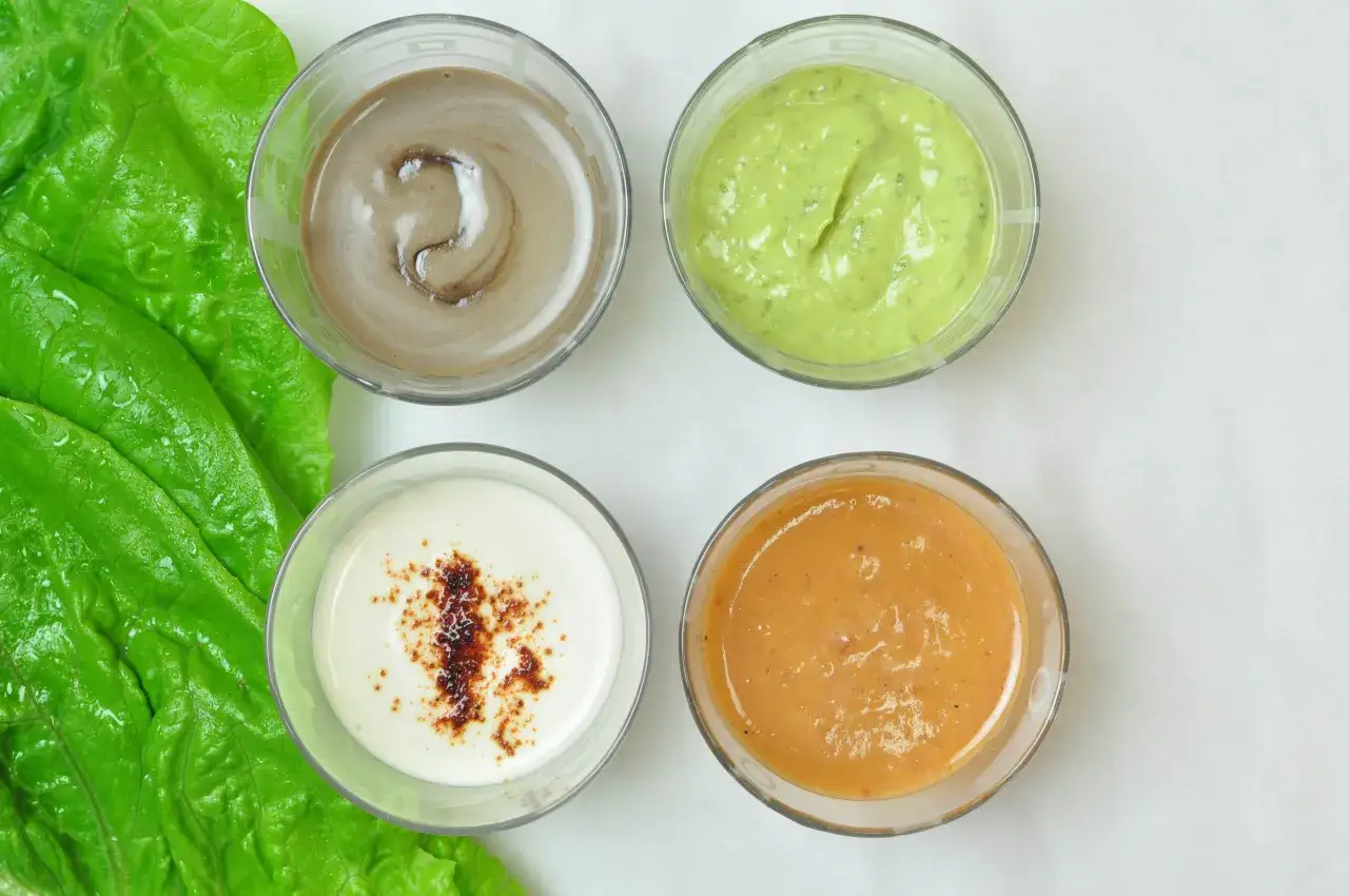 Fyra skålar med olika dressing till sallad, bredvid gröna salladsblad.