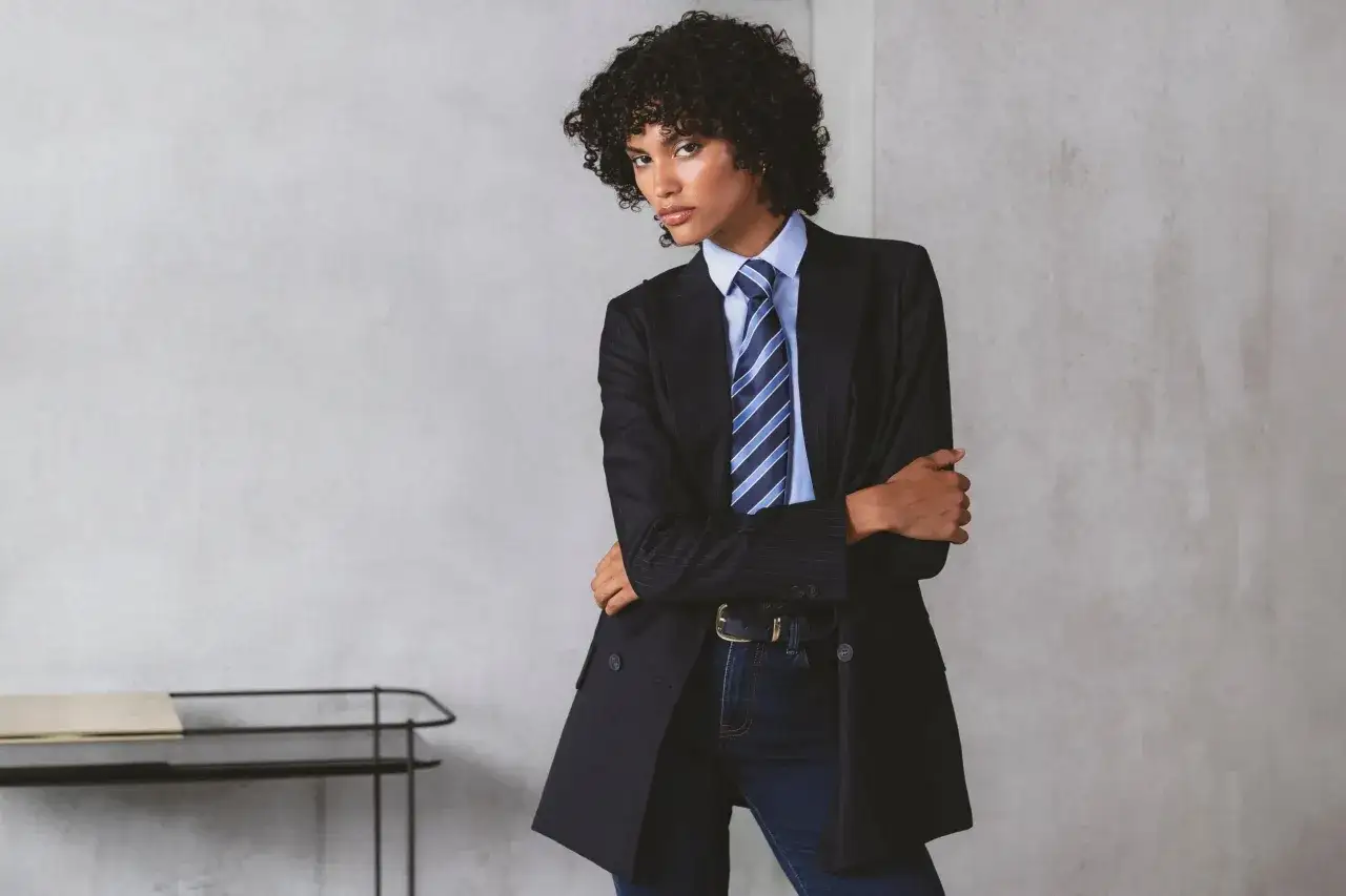 Una persona con rizos oscuros luce un traje a rayas, camisa azul y corbata. Su mirada y estilo transmiten una **belleza andr&oacute;gina** cautivadora.