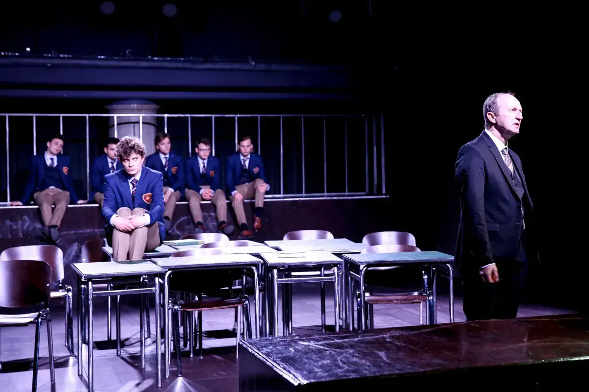 Chłopcy w mundurkach siedzą na ławce, a nauczyciel stoi przed nimi. Scena z teatru 