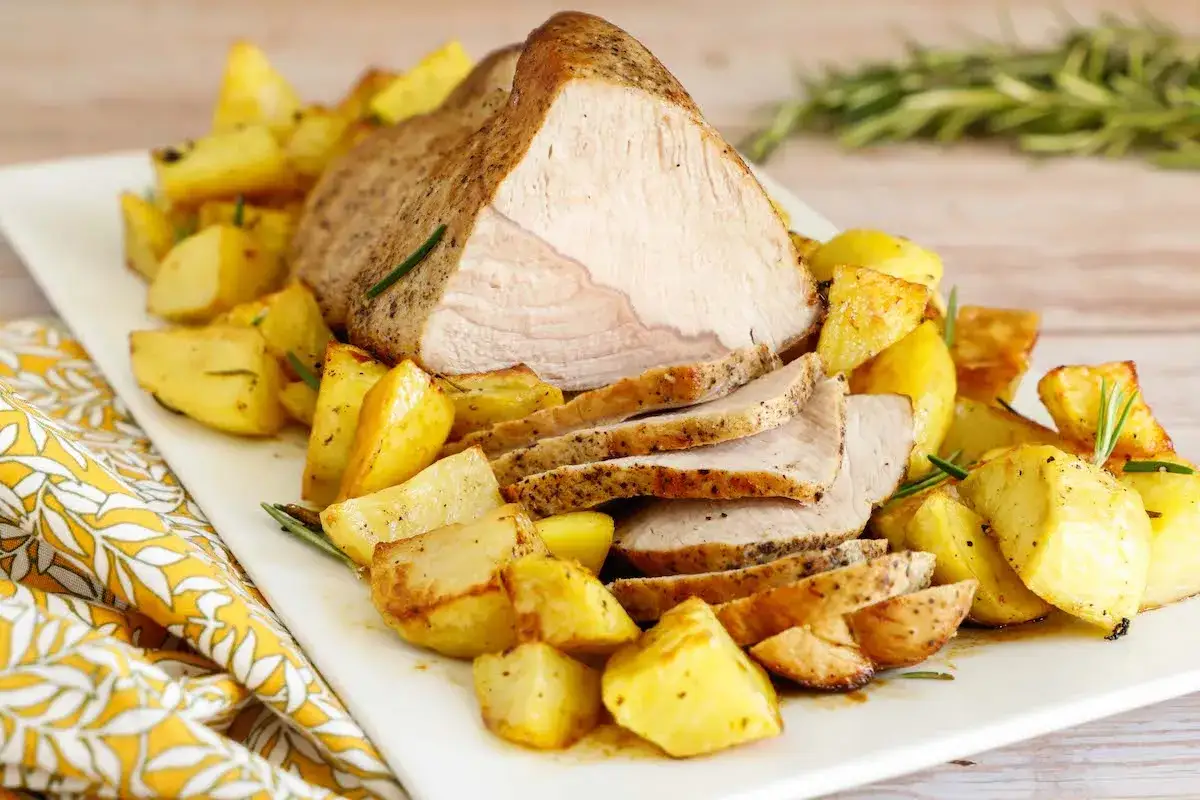 Un succulento arrosto di vitello affettato, circondato da patate arrosto dorate e profumate al rosmarino, servito su un piatto bianco.