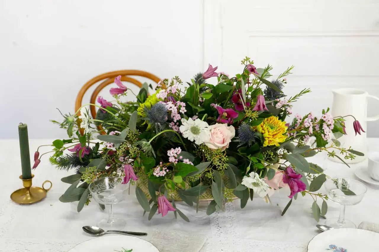 Un magnifique bouquet de fleurs sauvages, aux teintes roses, violettes et jaunes, est posé sur une table dressée. La mousse pour piquer des fleurs sert de base à cette composition florale.