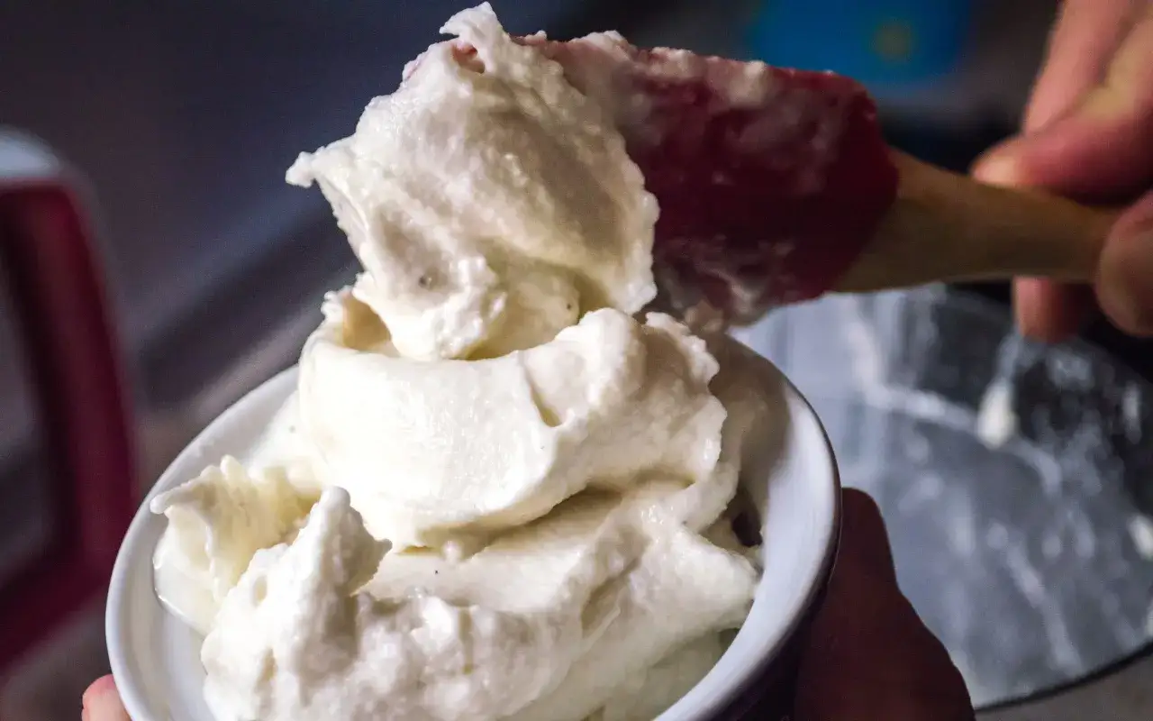 Un cucchiaio rosso preleva un'abbondante porzione di cremoso gelato fatto in casa senza gelatiera, servito in una ciotola bianca.