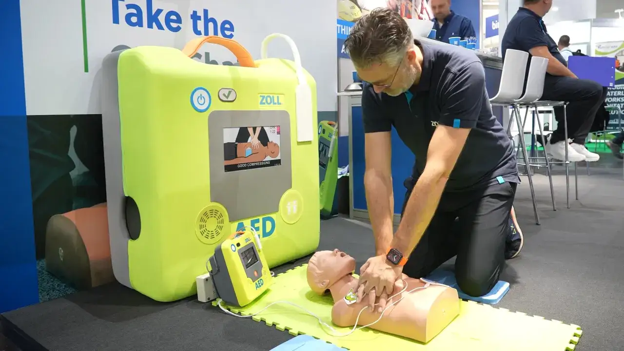 Mann &uuml;bt Reanimation an &Uuml;bungspuppe neben einem gro&szlig;en, gelben Defibrillator-Trainingsger&auml;t. Wann Defibrillator?