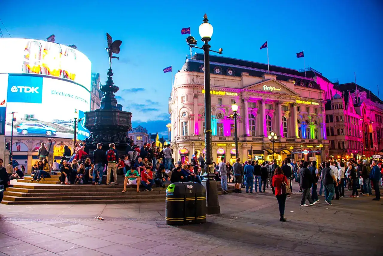 Tłum ludzi odpoczywa na schodach, podczas gdy neony rozświetlają budynki na Piccadilly Circus.