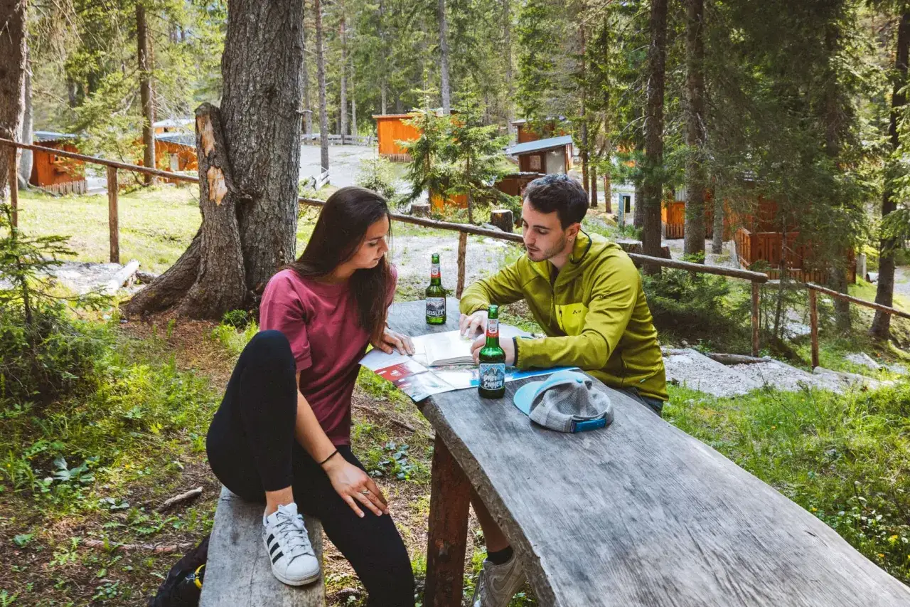 Para planują **camping** i analizują mapę przy piwie. W tle widać domki kempingowe.